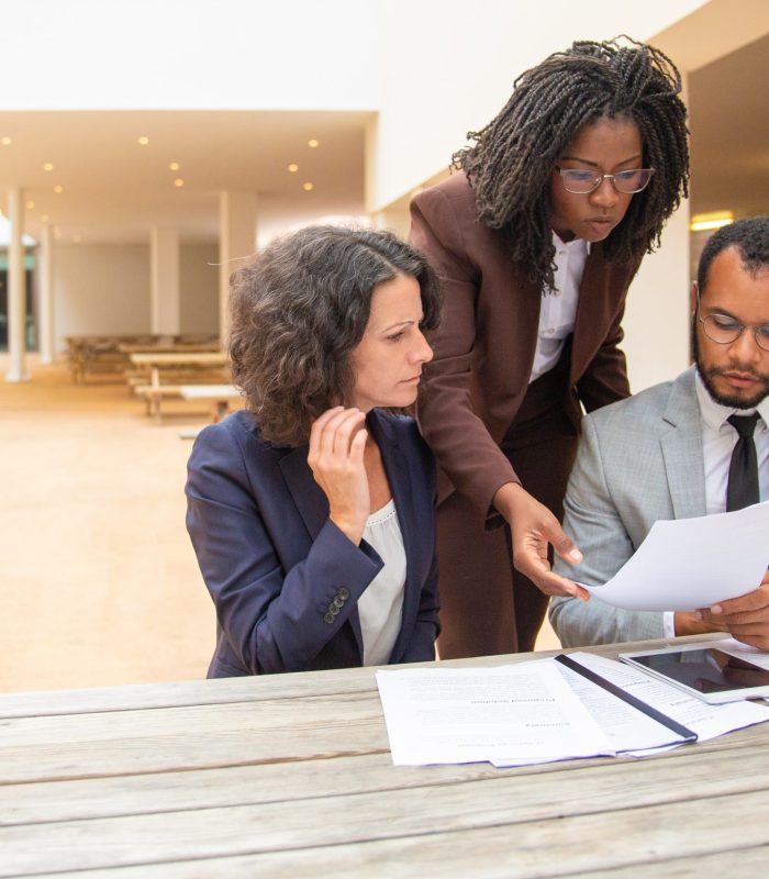 Business team of three studying documents in street cafe. Business man and women sitting and standing at table outside and staring at documents. Paper expertise concept